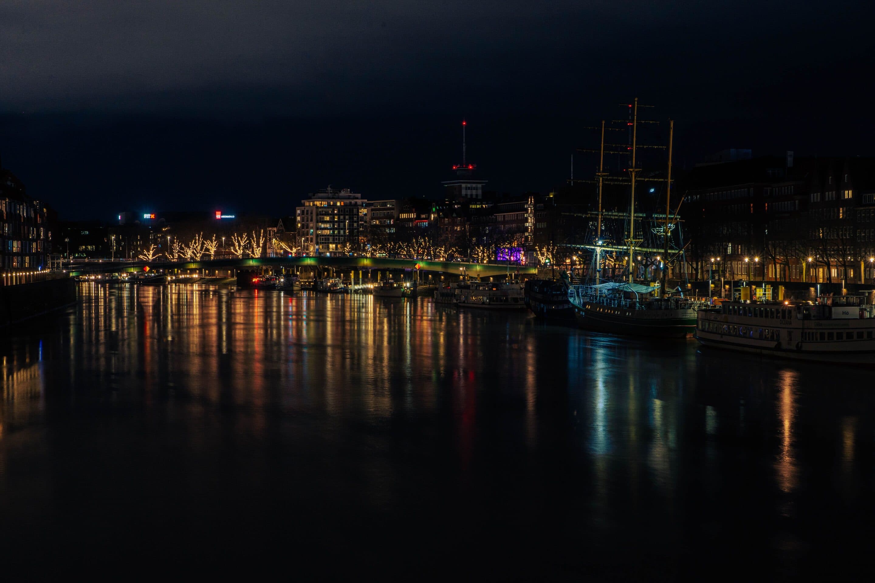 Die Bremer Schlachte bei Nacht: beleuchtete Uferbäume, Brücke und historische Segelschiffe an der Weser