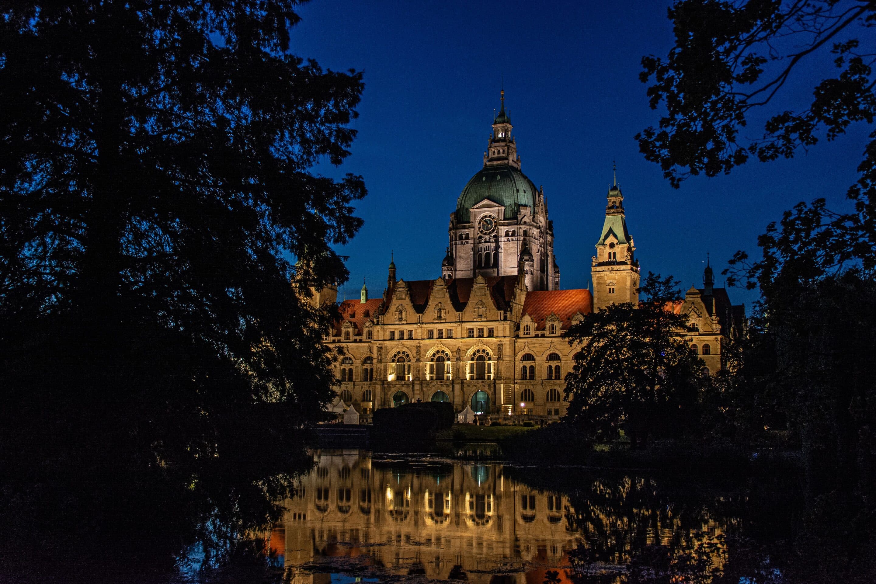 Das Neue Rathaus Hannover mit beleuchteter Kuppel und Türmen bei Nacht, gespiegelt im Wasser davor