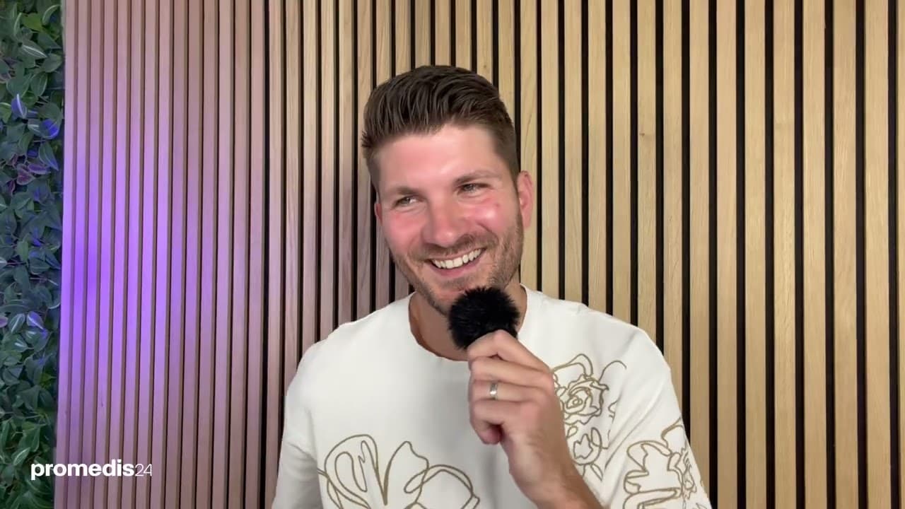 Moderator lächelt breit mit Mikrofon vor violett beleuchteter Holzlamellenwand – Fachkräfte erzählen von ihrem Berufsweg in der Zeitarbeit