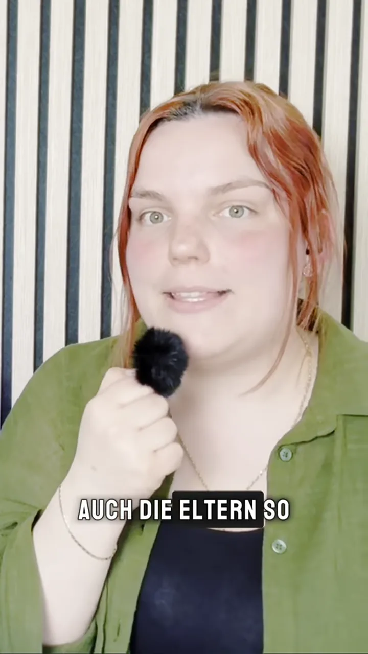 Eine Frau mit rotem Haar lacht mit Mikrofon vor einer Holzlattenwand, Untertitel im Bild