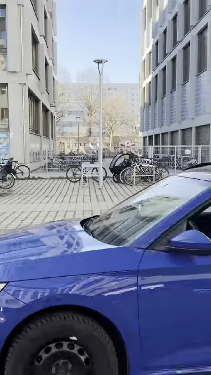 Eine Frau mit Sonnenbrille sitzt am Steuer eines blauen Autos zwischen Bürogebäuden, Untertitel im Bild