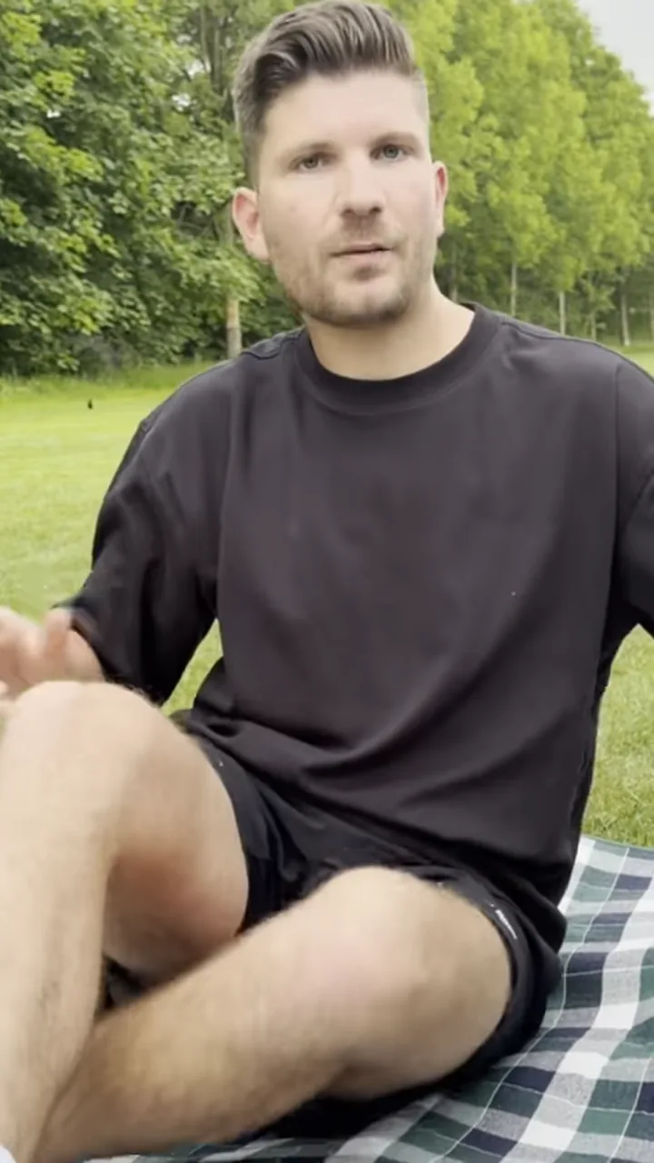 Ein Mann sitzt im Schneidersitz auf einer karierten Picknickdecke im Park und schaut in die Kamera