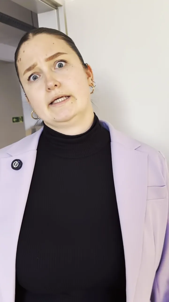 Eine Frau im fliederfarbenen Blazer steht in einem Flur und spricht in die Kamera