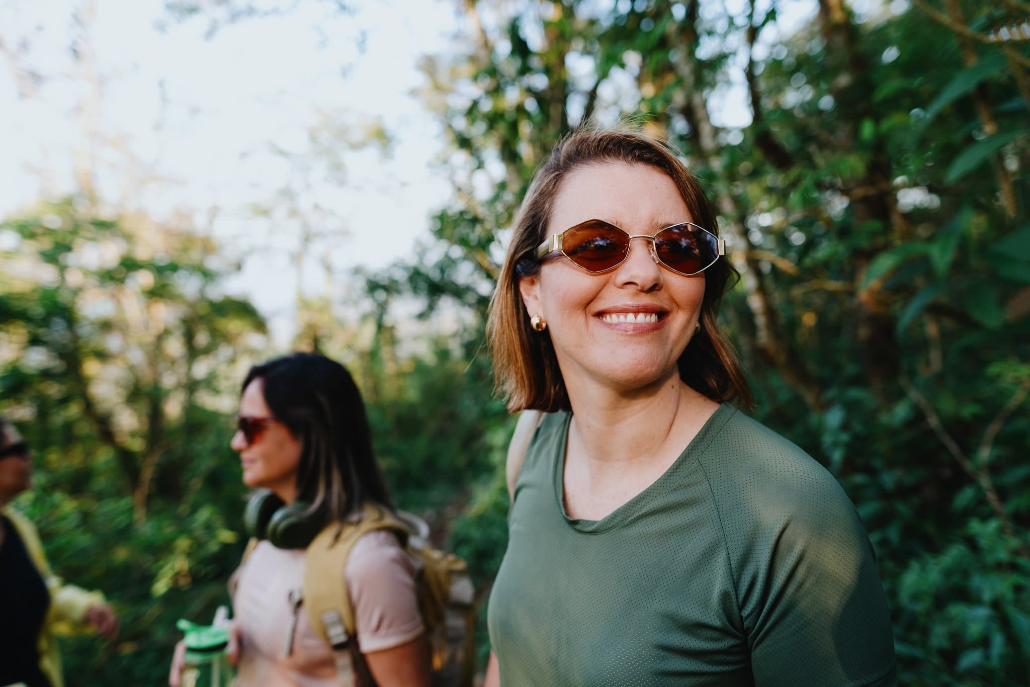 Frau mit Sonnenbrille lächelt bei einer Wanderung im Wald, hinter ihr eine Begleiterin