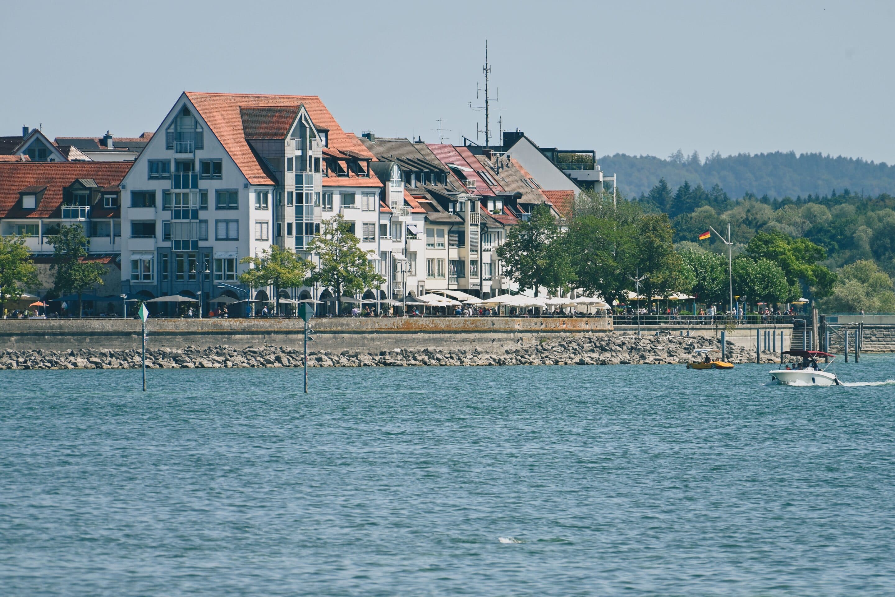Die Uferpromenade von Friedrichshafen am Bodensee mit Booten, Cafés und bewaldeten Hügeln im Hintergrund